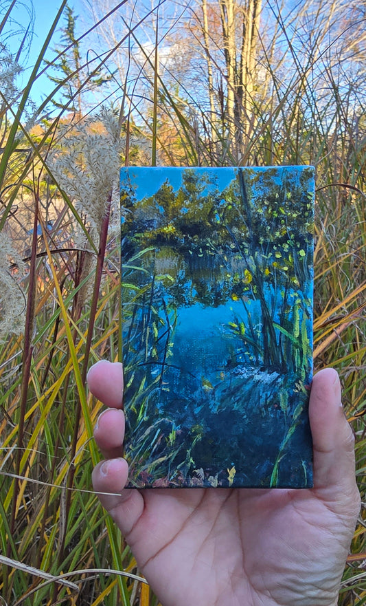 Hand holding a small rectangular painting of a landscape with trees and water, surrounded by natural elements.