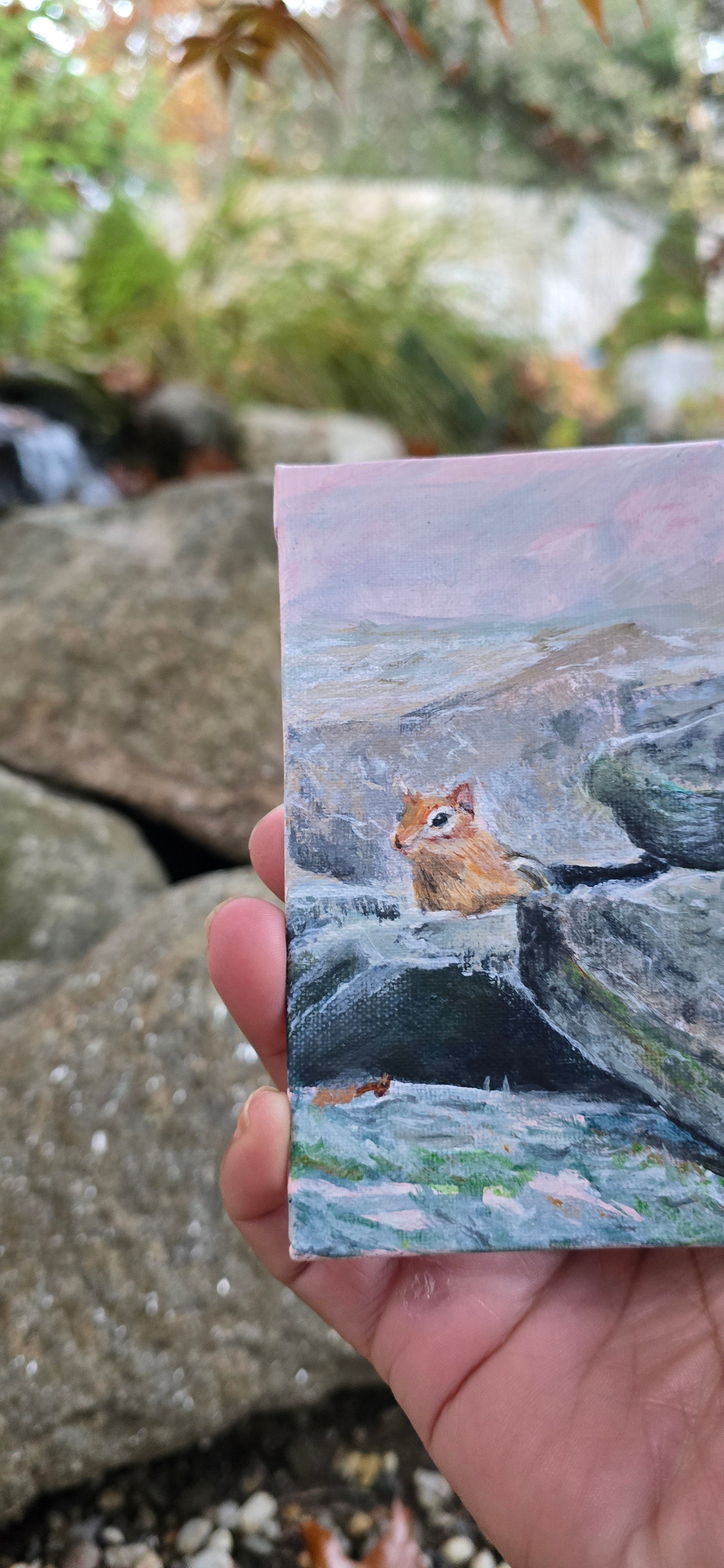 Hand holding a small painting of a chipmonk on a rock with a natural background.