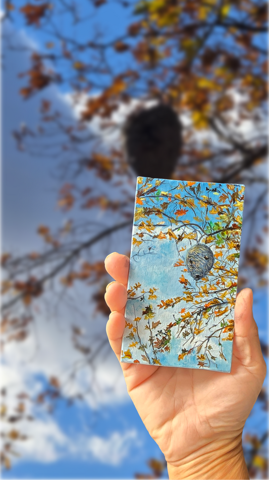 Hand holding a small painting with autumn design against a blue sky with tree branches.