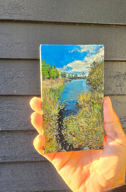 Hand holding a small rectangular painting of a landscape with blue sky and green grass.